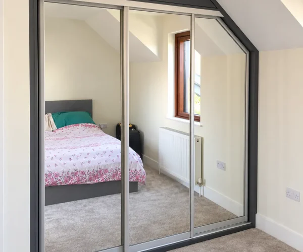 Bespoke mirrored sliding wardrobe fitted beneath a sloping attic ceiling with a silver frame and full-length mirrors.